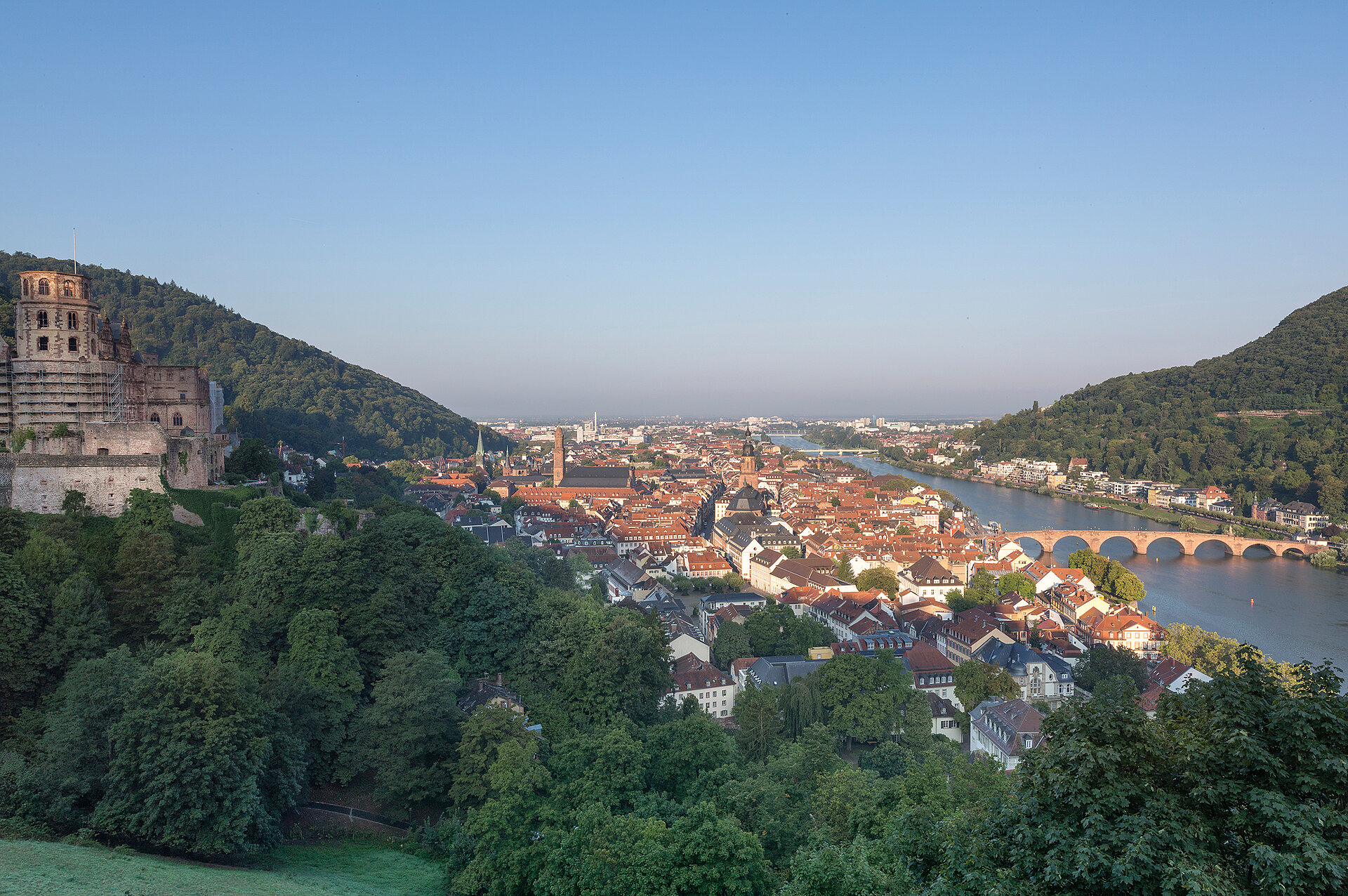 Blick über eine historische Stadt am Fluss mit roter Dachlandschaft, Brücke und grünen Hügeln; links eine alte Burganlage.