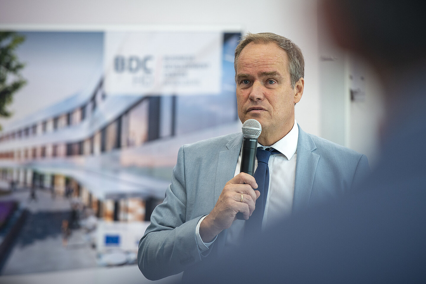 Ein Mann im hellen Anzug spricht mit einem Mikrofon bei einer Veranstaltung vor einem unscharfen Hintergrund.