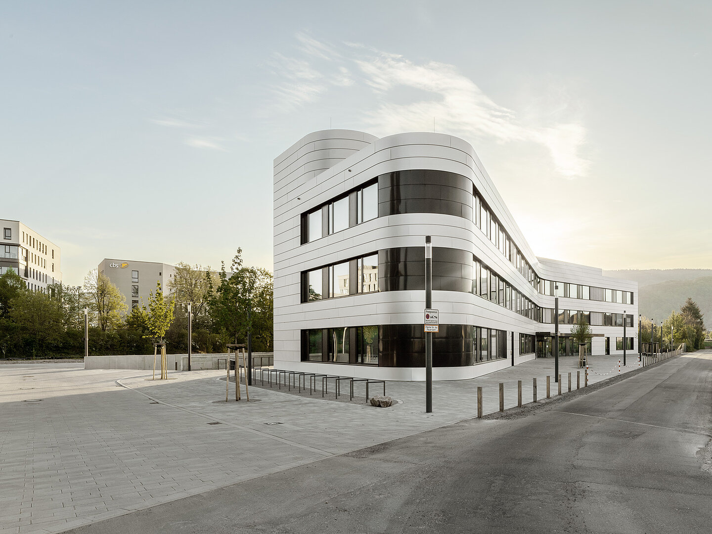 Modernes, weißes Bürogebäude mit geschwungener Fassade an einer leeren Straße, umgeben von einem offenen Vorplatz und wenigen Bäumen.
