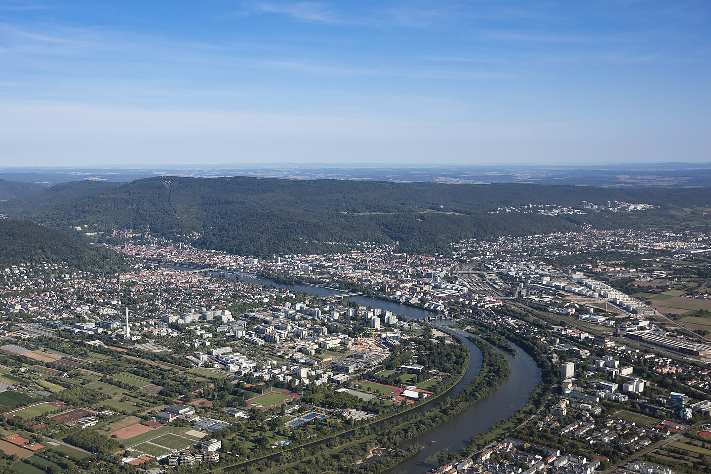 Luftaufnahme einer Stadt mit einem mäandrierenden Fluss, umgeben von Wäldern, Feldern und Hügeln unter klarem blauem Himmel.