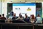 Ein Mann hält einen Vortrag vor sitzendem Publikum in einem modernen Konferenzraum; auf der Leinwand sind Logos von Sponsoren oder Partnern zu sehen.