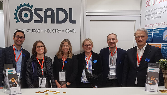 Sechs Personen in Businesskleidung stehen an einem Messestand von OSADL und lächeln in die Kamera.