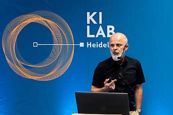 Ein Mann hält einen Vortrag vor einem Laptop auf einer Bühne mit dem Schriftzug „KI LAB Heidelberg“ und einer grafischen Kreisillustration im Hintergrund.