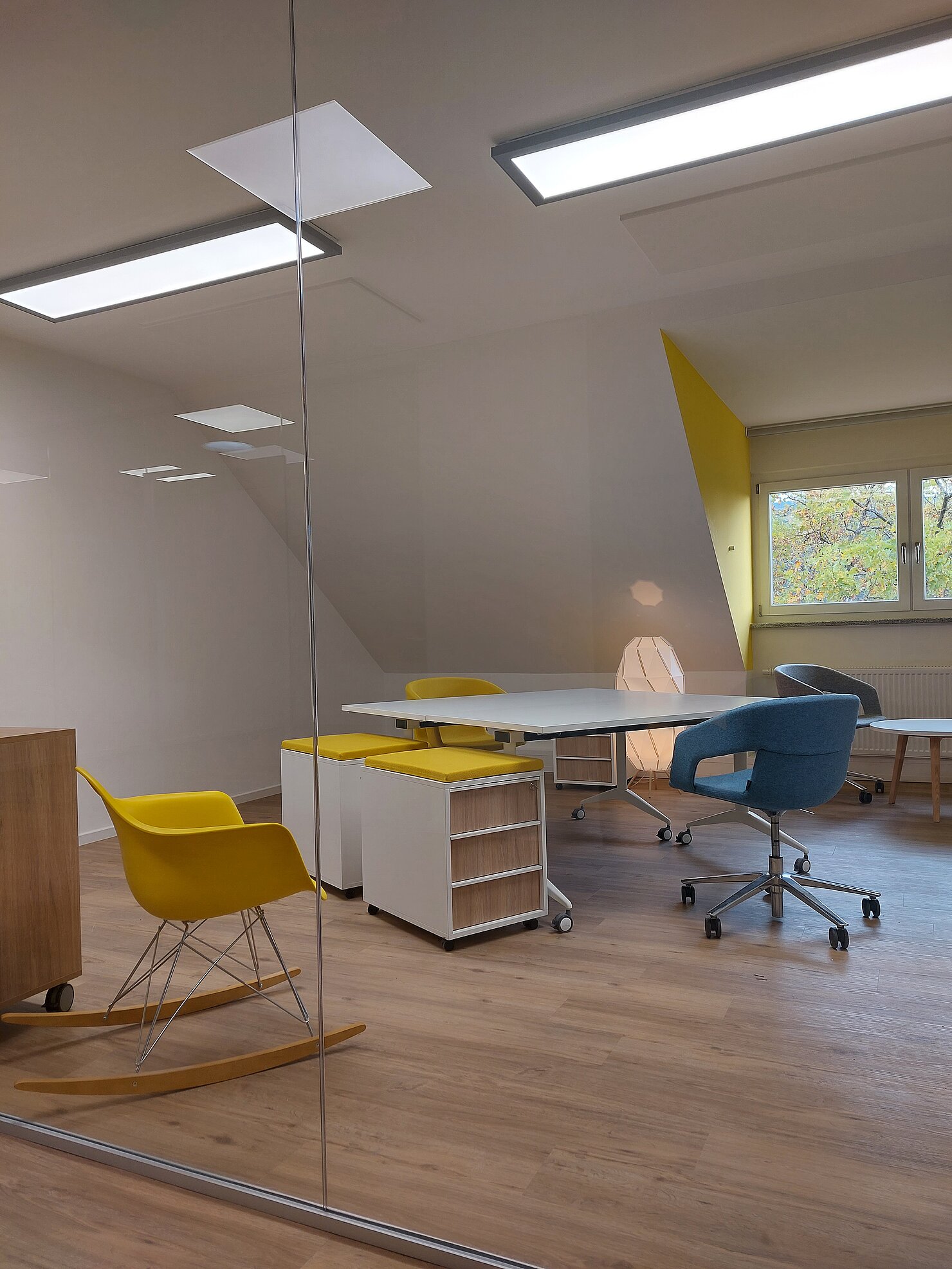 Modernes Büro mit hellen Holzböden, großen Fenstern und mehreren Schreibtischen sowie bunten Stühlen in Gelb und Blau.