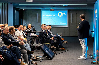 Ein Mann hält einen Vortrag vor sitzendem Publikum in einem Konferenzraum mit Präsentationsleinwand.