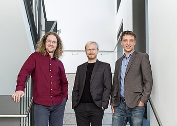Drei Männer stehen lächelnd auf einer Treppe in einem modernen Gebäude.