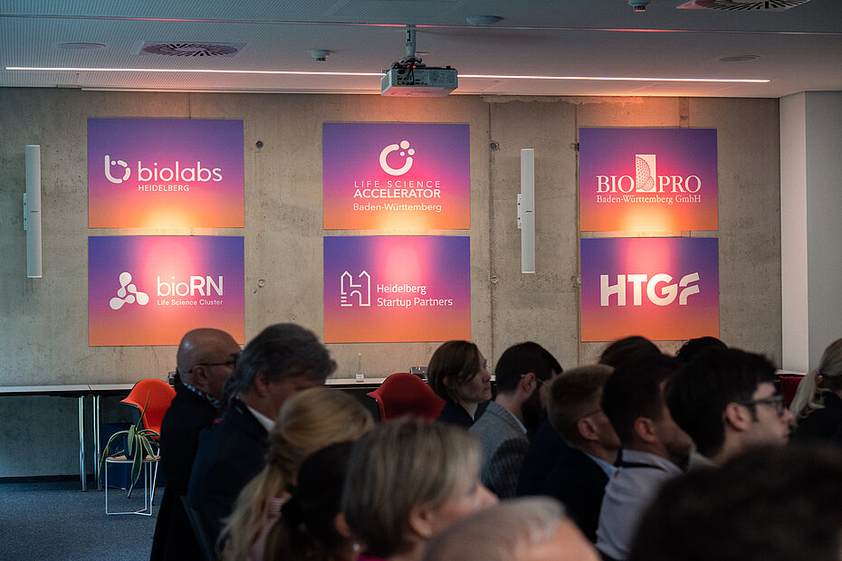 Publikum sitzt in einem Konferenzraum vor einer Wand mit leuchtenden Logos von Biolabs Heidelberg, Life Science Accelerator Baden-Württemberg, BioPRO, bioRN, Heidelberg Startup Partners und HTGF.