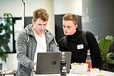 Zwei Männer stehen an einem Tisch und schauen gemeinsam auf einen Laptop, offenbar bei einer Besprechung oder Präsentation.