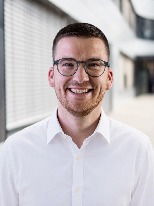 Lächelnder Mann mit Brille und weißem Hemd in einem hellen Innenraum.