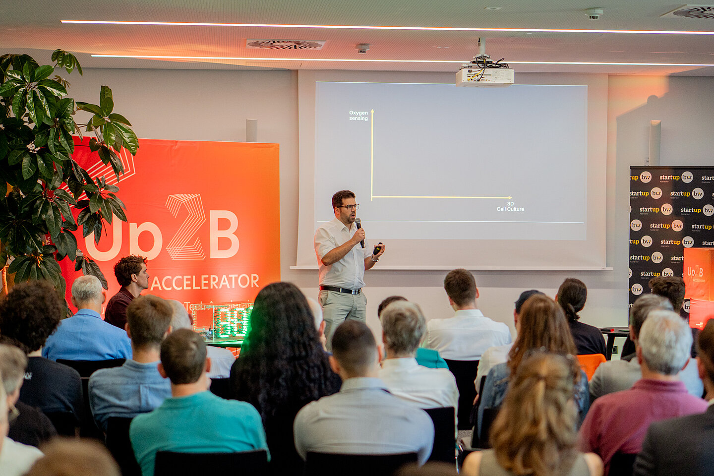 Ein Mann hält bei einem Vortrag ein Mikrofon und präsentiert vor sitzendem Publikum eine Folie auf einer Leinwand in einem Konferenzraum.