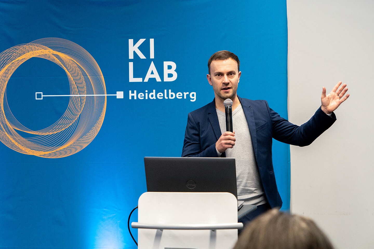 Ein Mann hält an einem Podium mit Laptop ein Mikrofon und spricht vor einer KI-LAB-Heidelberg-Kulisse, während er mit der anderen Hand gestikuliert.