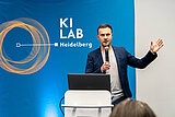 Ein Mann hält an einem Podium mit Laptop ein Mikrofon und spricht vor einer KI-LAB-Heidelberg-Kulisse, während er mit der anderen Hand gestikuliert.