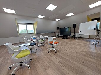 Heller Schulungsraum mit mehreren modernen Stühlen, Tischen, einem großen Bildschirm und einem Whiteboard.